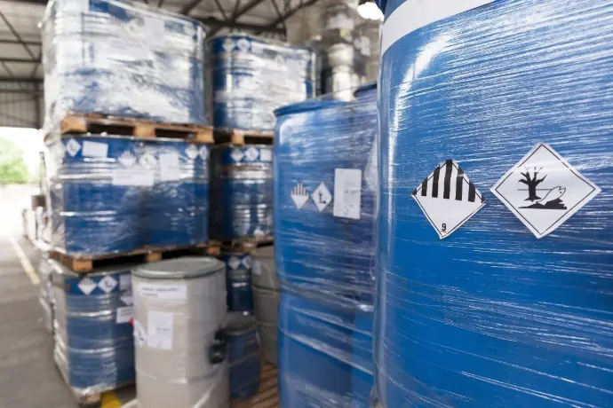Pallets of blue industrial chemical drums with hazard labels stored in a warehouse.